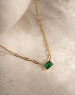 Gold paperclip necklace with a emerald pendant gemstone on a beige marble surface