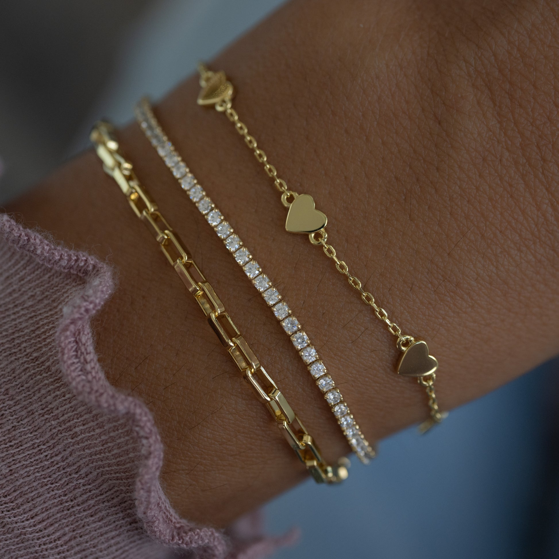 model wearing  layered bracelets featuring a gold heart charm bracelet styled with a paperclip bracelet and a tennis bracelet