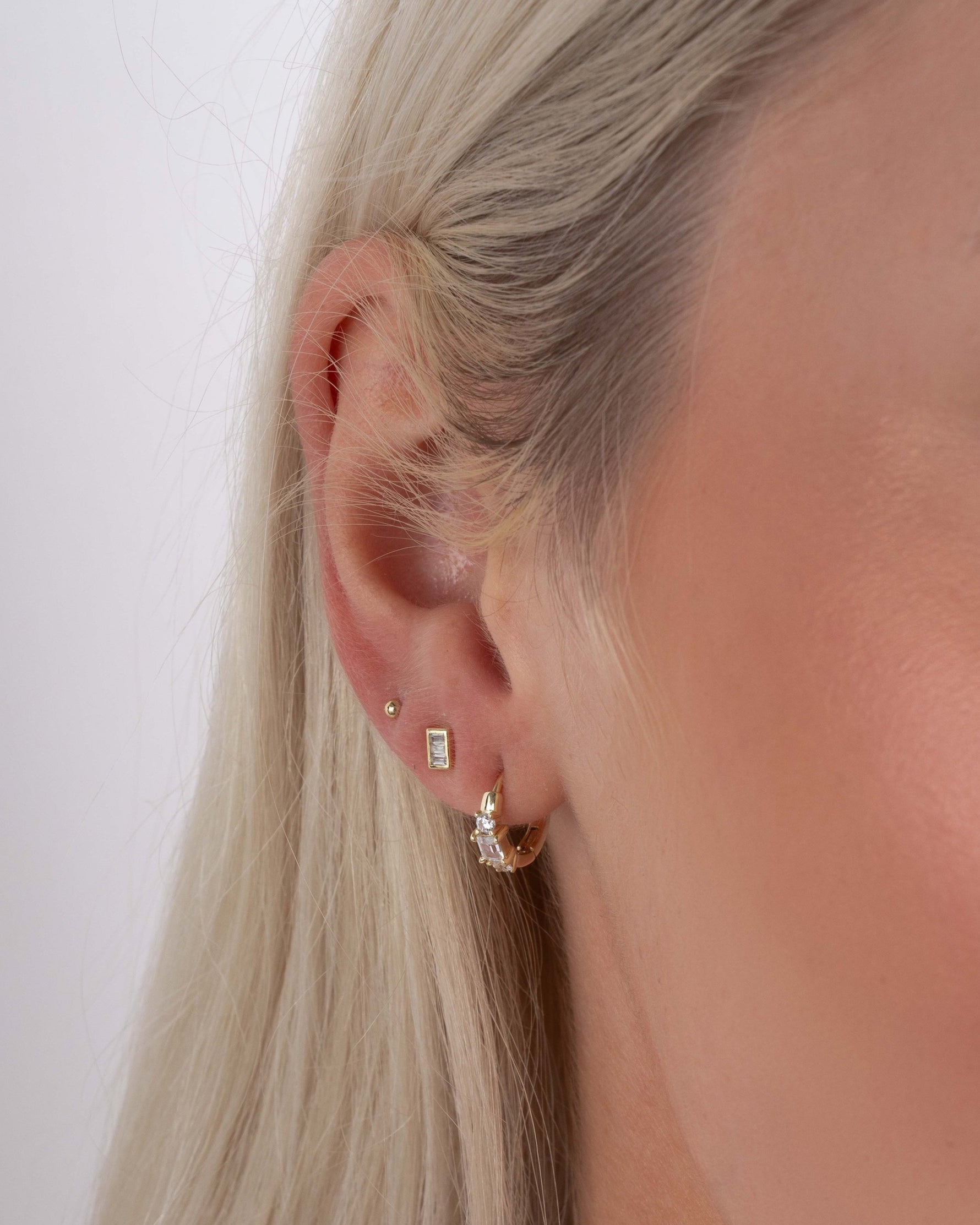 Close-up of a model wearing three gold earrings including a pair of dainty huggie hoops, minimalist stud earrings, and small ball studs