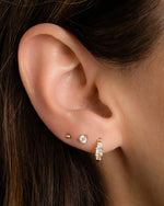 closeup image of gold earring stack with cz three stone huggie hoop, a dainty stud earring, and a small ball stud