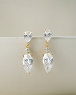 Bridal earrings featuring gold stud earrings with clear teardrop-shaped gemstones on a beige background