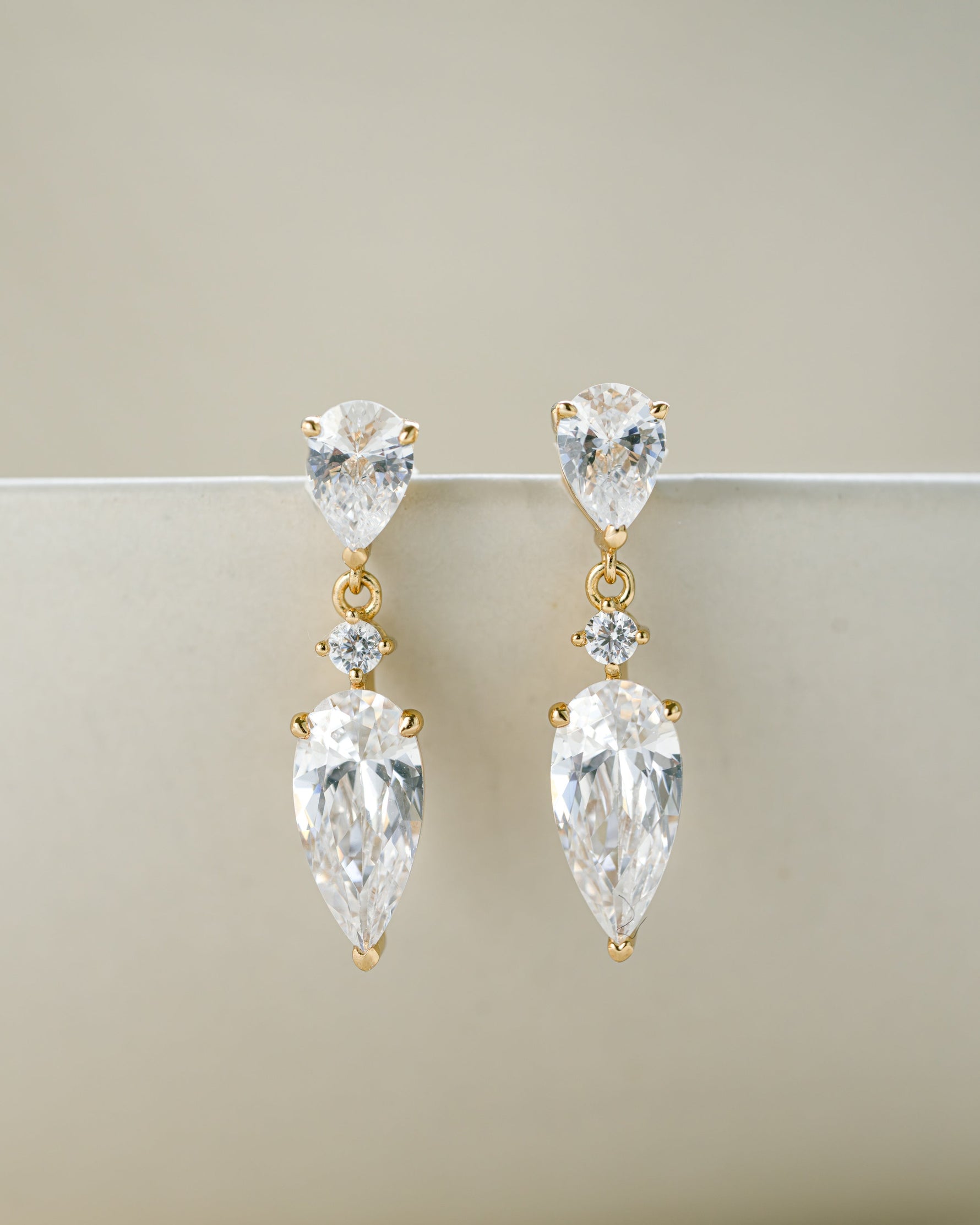 Bridal earrings featuring gold stud earrings with clear teardrop-shaped gemstones on a beige background