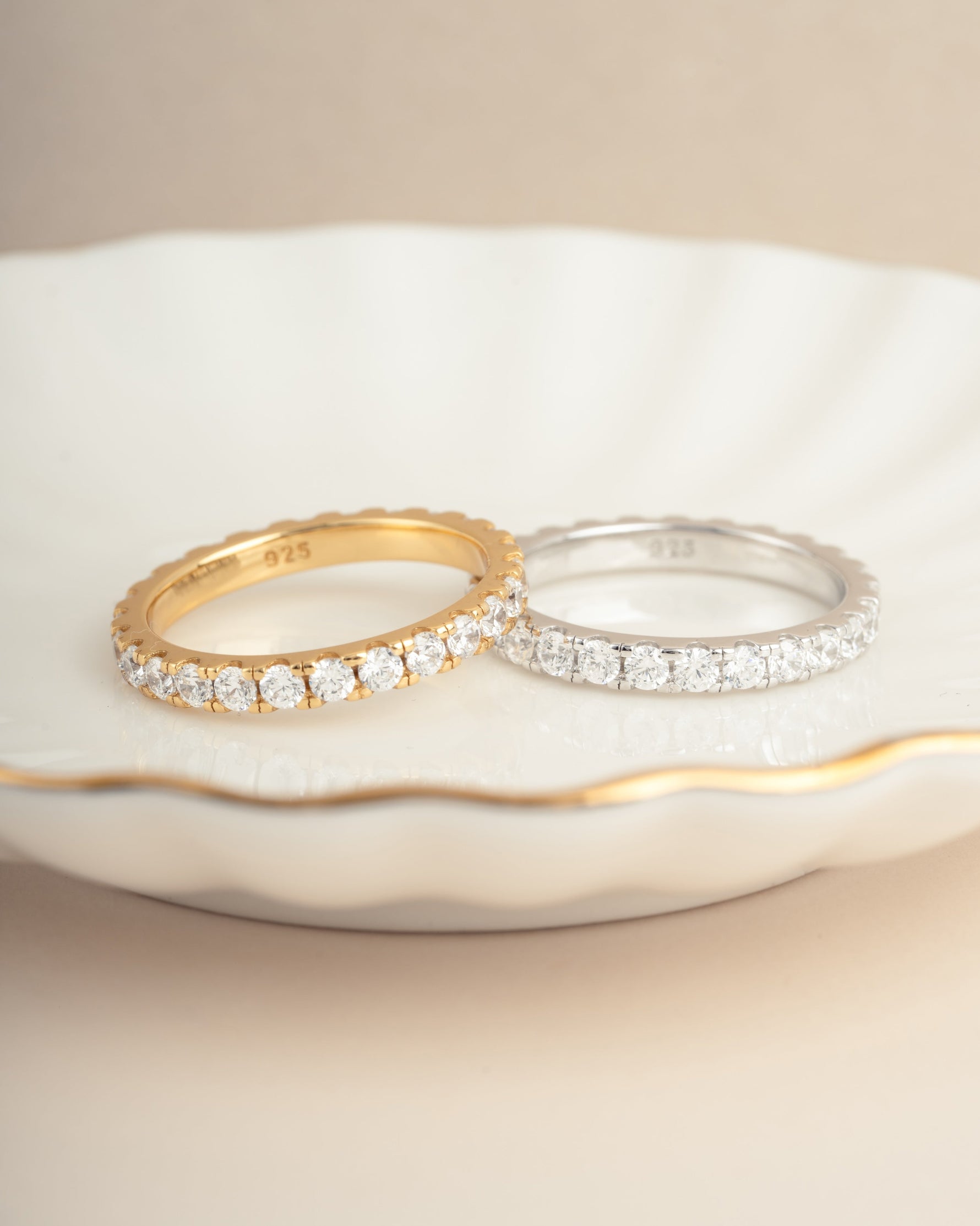 Two diamond rings lying next to eachother on a decorative plate, one gold wedding band with cubic zirconia stones, and one sterling silver dainty ring with cubic zirconia stones