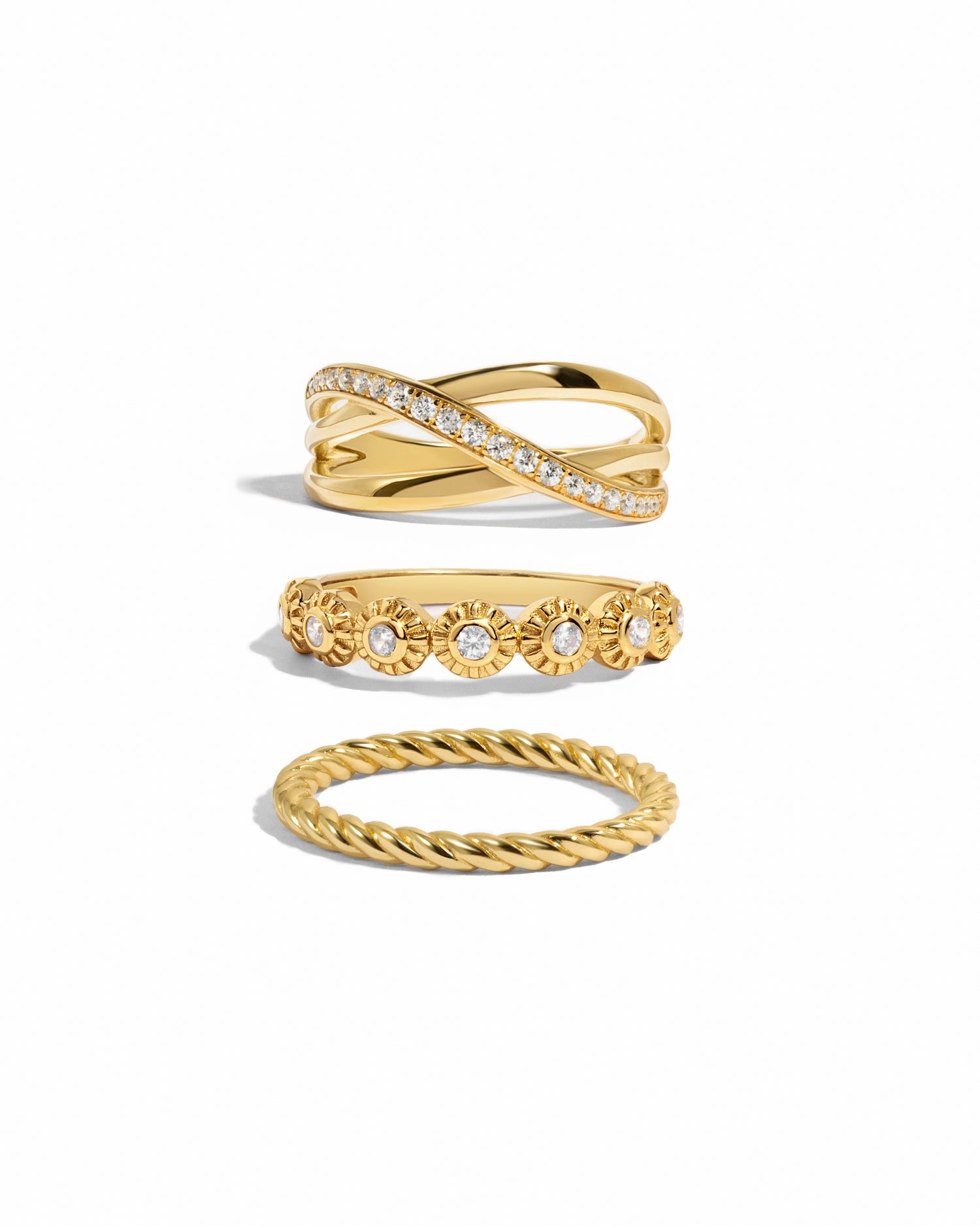 Gold stackable rings including a diamond x band ring with a diamond slash, a gold bezel set textured gold ring, and a thick spiral band ring on a white background