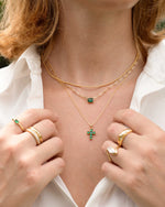 Closeup image of a minimalist layering necklaces featuring an emerald pendant necklace, a cross necklace, and a gold chain necklace
