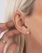 Model wearing gold tiny earrings and gold stackable rings, featuring a gold ball stud earring, a dainty diamond earring, and a gold flower earring 