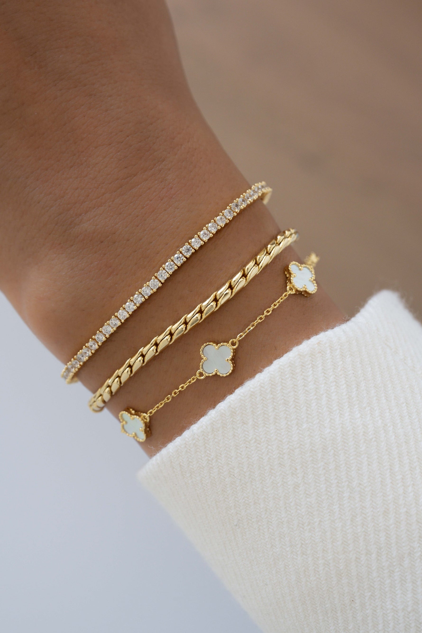 A close-up of a person's wrist wearing four delicate gold bracelets: a tennis bracelet with clear stones, a twisted rope chain bracelet, and two gold chain bracelets with white flower charms.