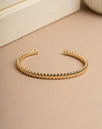 Gold cuff with a rope bracelet design on a beige surface with a book in the background