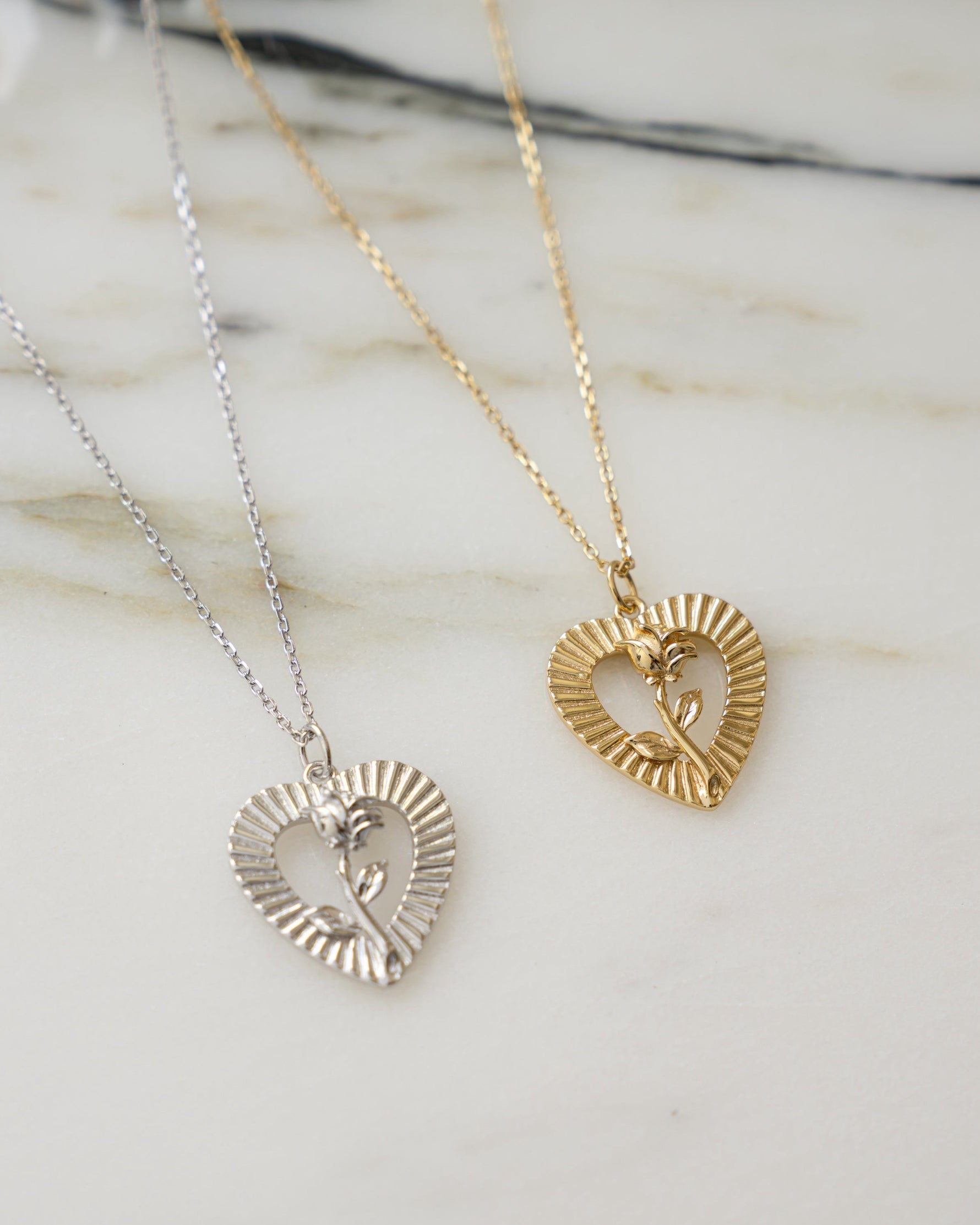 Two chain necklaces on a marble background featuring a sterling silver chain necklace with a heart pendant  and a gold statement necklace with a rose pendant  