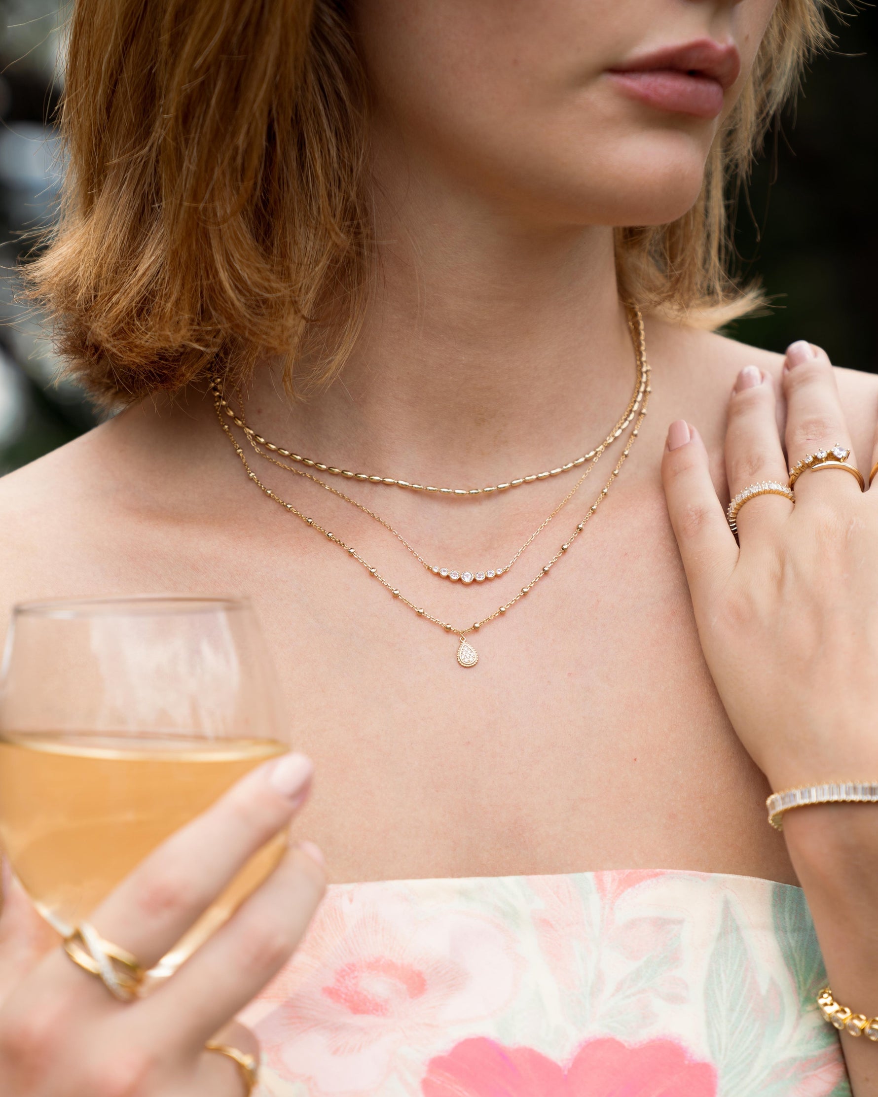 Woman holding a glass of wine with stunning gold jewelry featuring gold layering necklaces, one simple beaded necklace, a gold gradient diamond bezel set bar necklace, and a beaded charm necklace, paired with dainty gold stackable rings 
