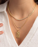 Gold layering necklaces on a person wearing a white sweater featuring a round bezel tennis necklace, a dainty gold bar necklace, and a rectangle rose medallion necklace 
