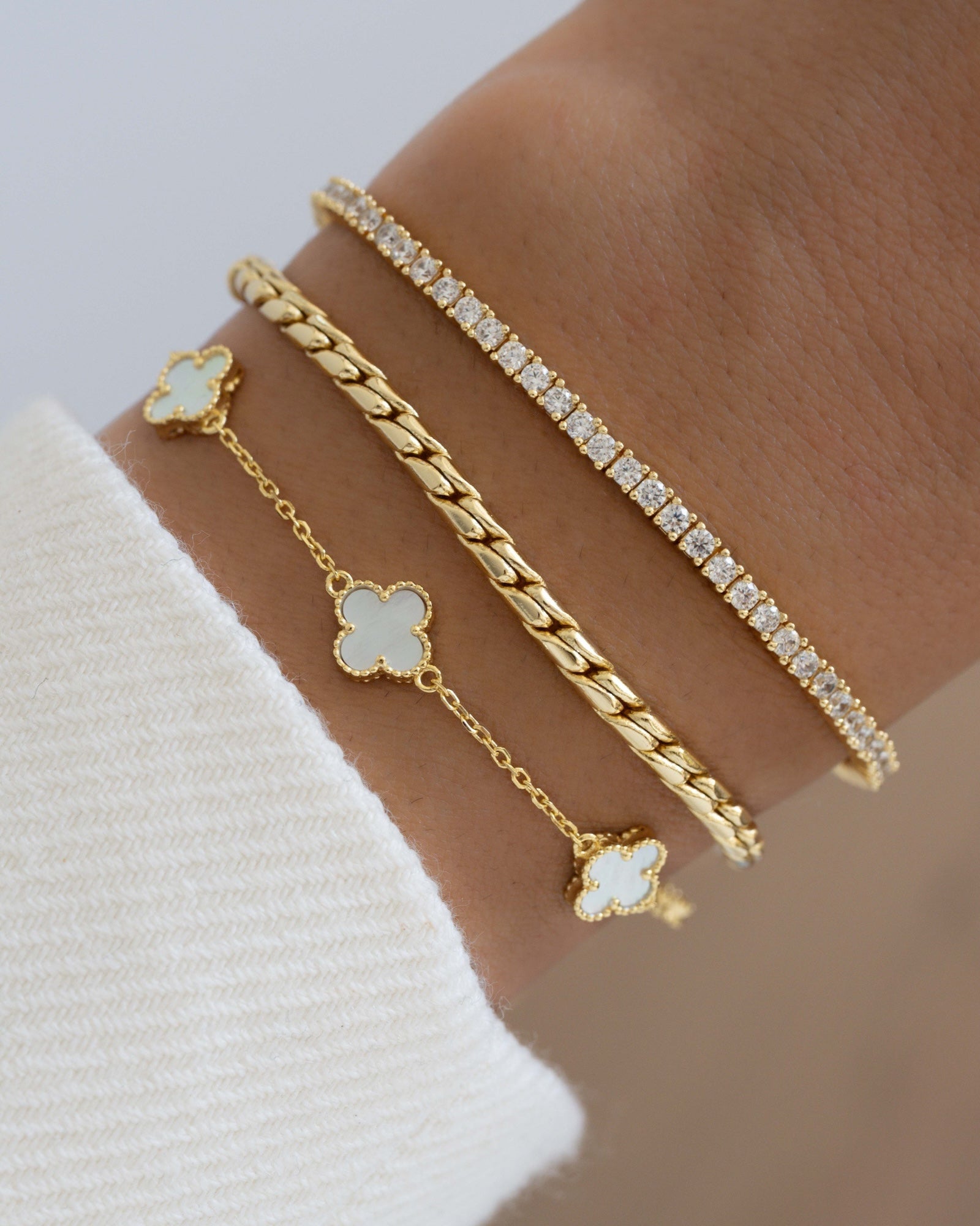 Gold stackable bracelets, featuring a pearl four leaf clover bracelet, a chunky gold chain bracelet, and a minimalist gold tennis bracelet 