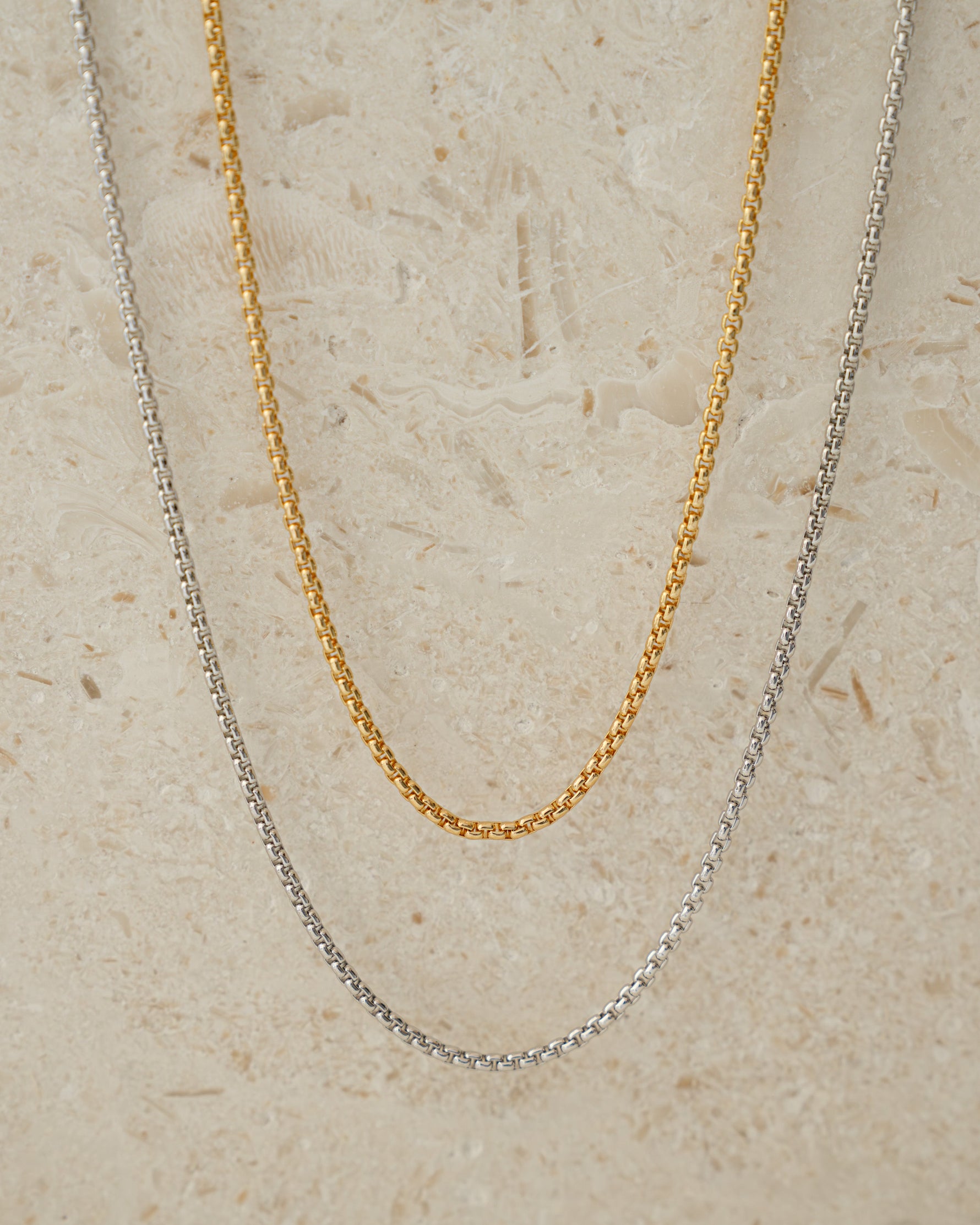 Two gold and silver chain necklaces on a beige textured background