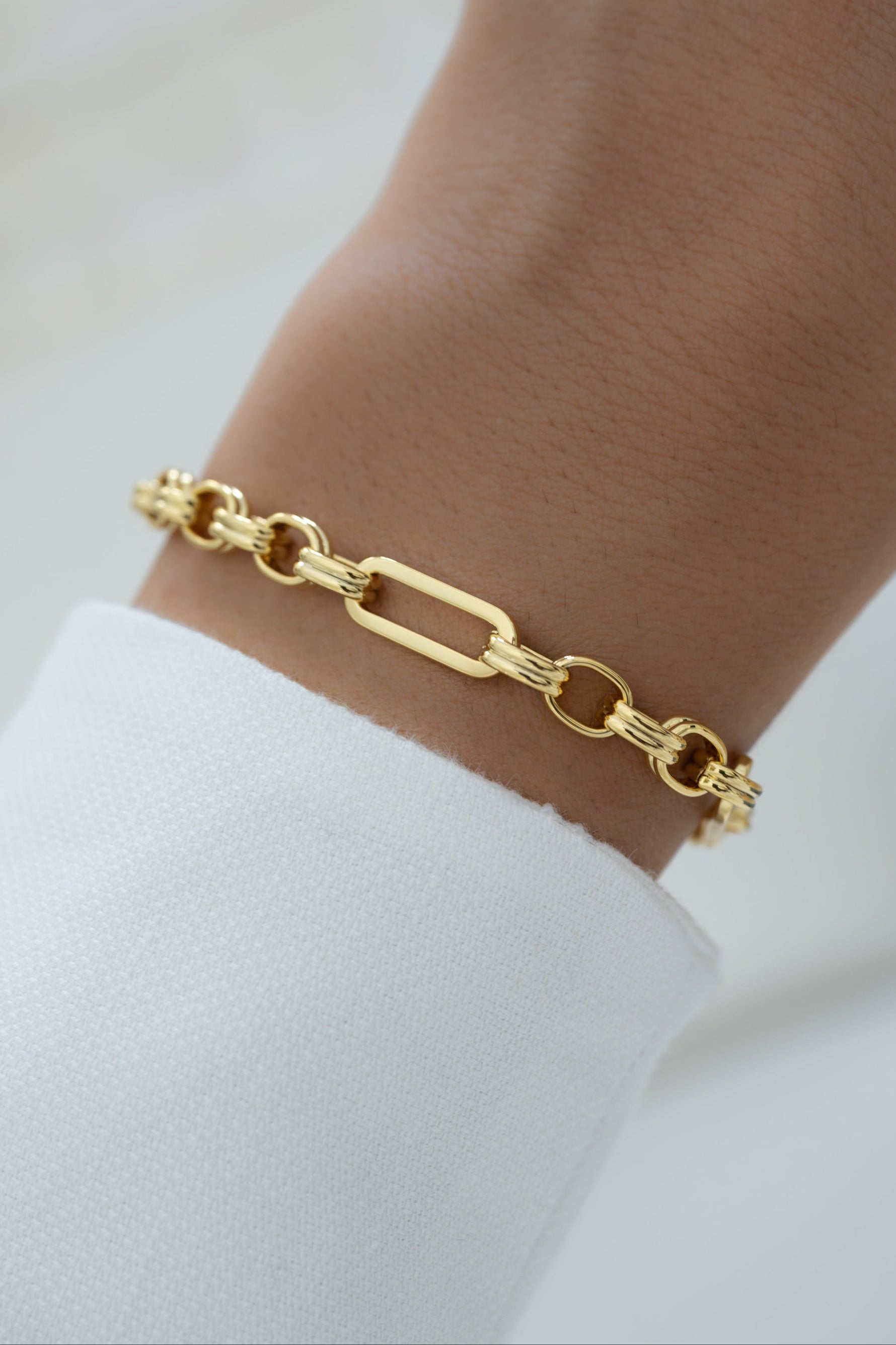 A gold paperclip chain link bracelet worn on a person's wrist, shown in a close-up shot with a white sleeve visible.