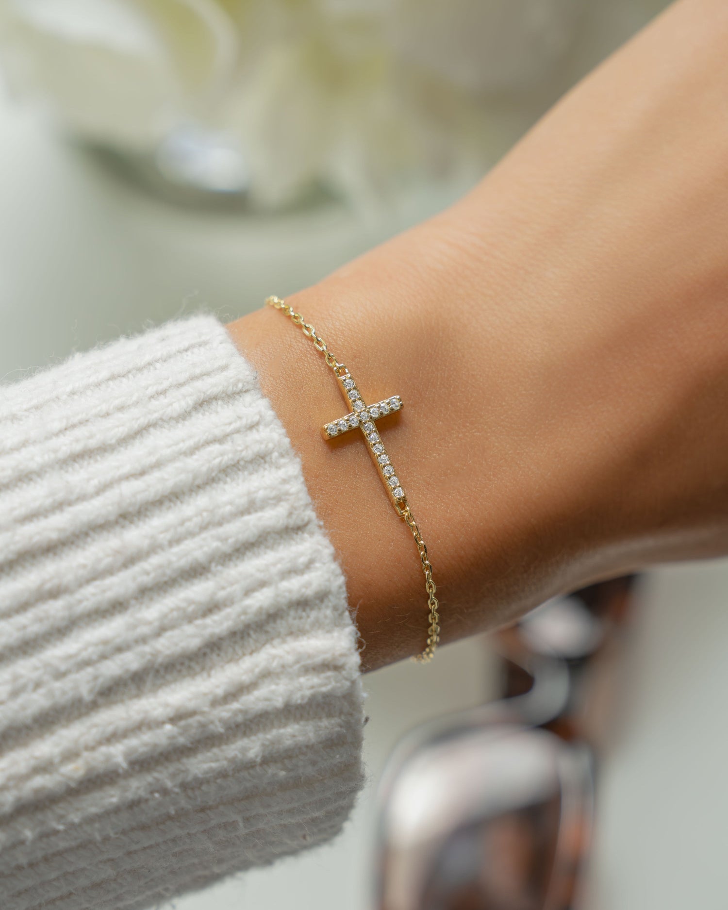 Gold chain bracelet with a cross bracelet design on a wrist, blurred background with flowers