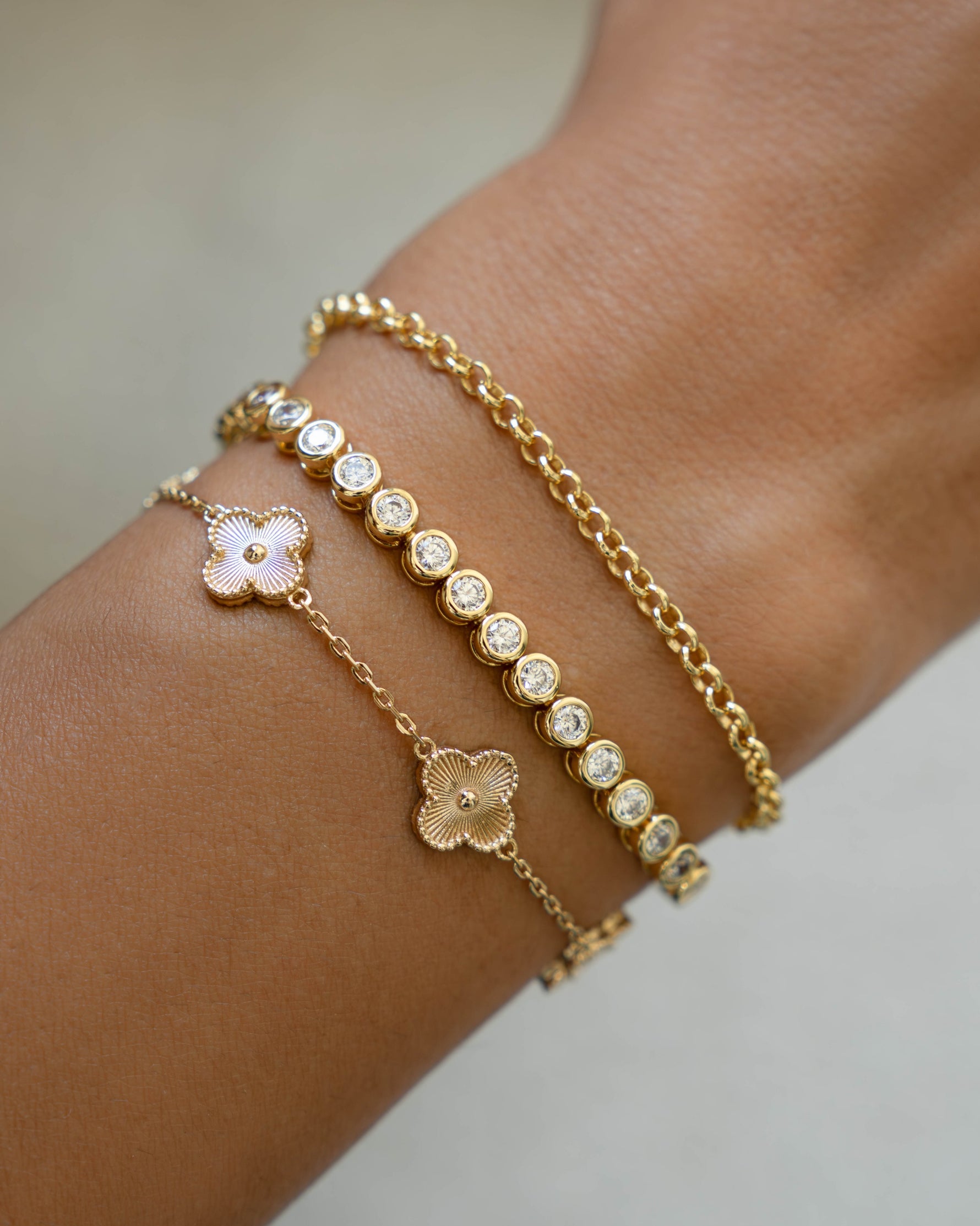 Gold bracelet  stack with four leaf clover design, a simple round bezel tennis bracelet, and a dainty gold chain bracelet on a wrist against a neutral background