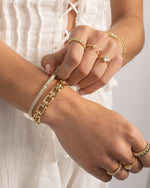 Closeup of a model wearing gold jewelry featuring gold stackable rings, a gold tennis bracelet, and a paperclip chain statement bracelet 