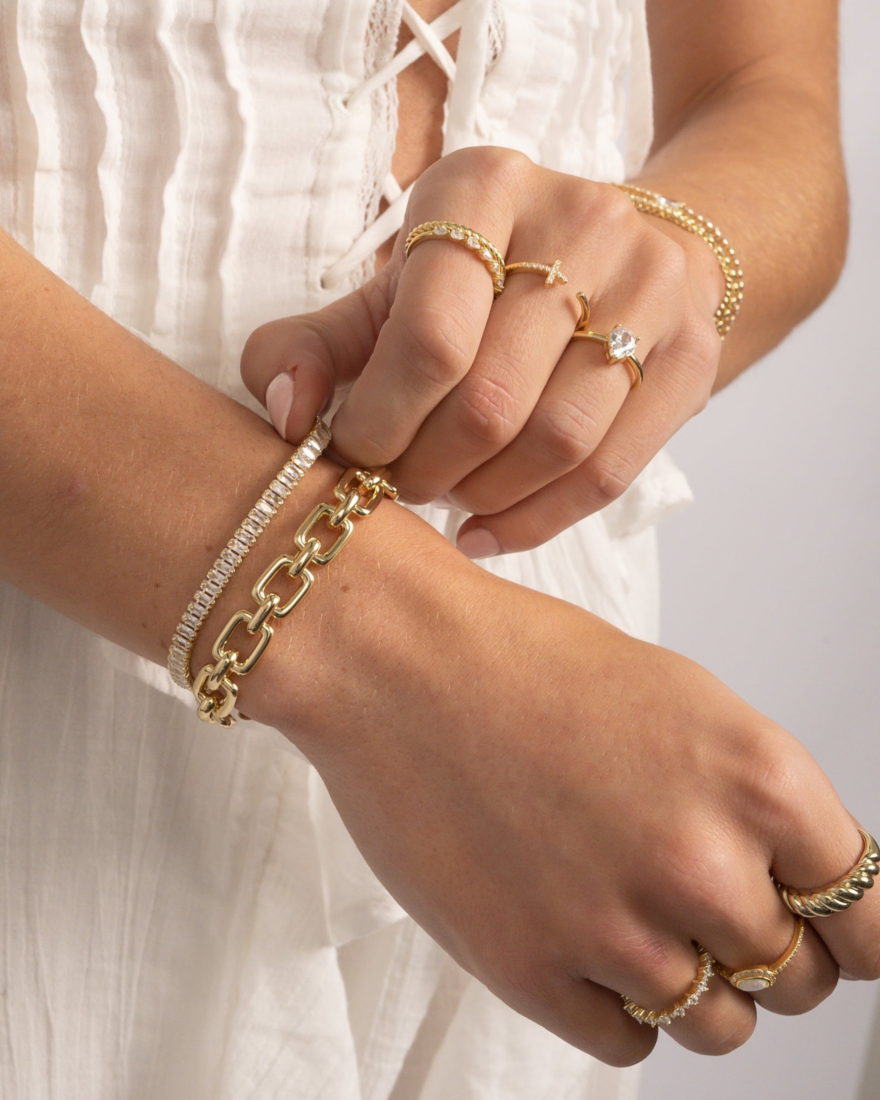 Closeup of a model wearing gold jewelry featuring gold stackable rings, a gold tennis bracelet, and a paperclip chain statement bracelet 