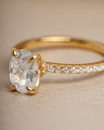 Gold oval engagement ring with a pave diamond band on a beige background