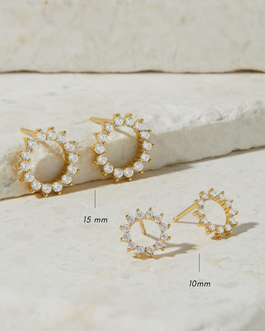 Gold diamond stud earrings in 10mm and 15mm on a textured beige surface