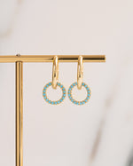 Small gold charm hoop earrings with turquoise gemstones on a gold stand against a light background