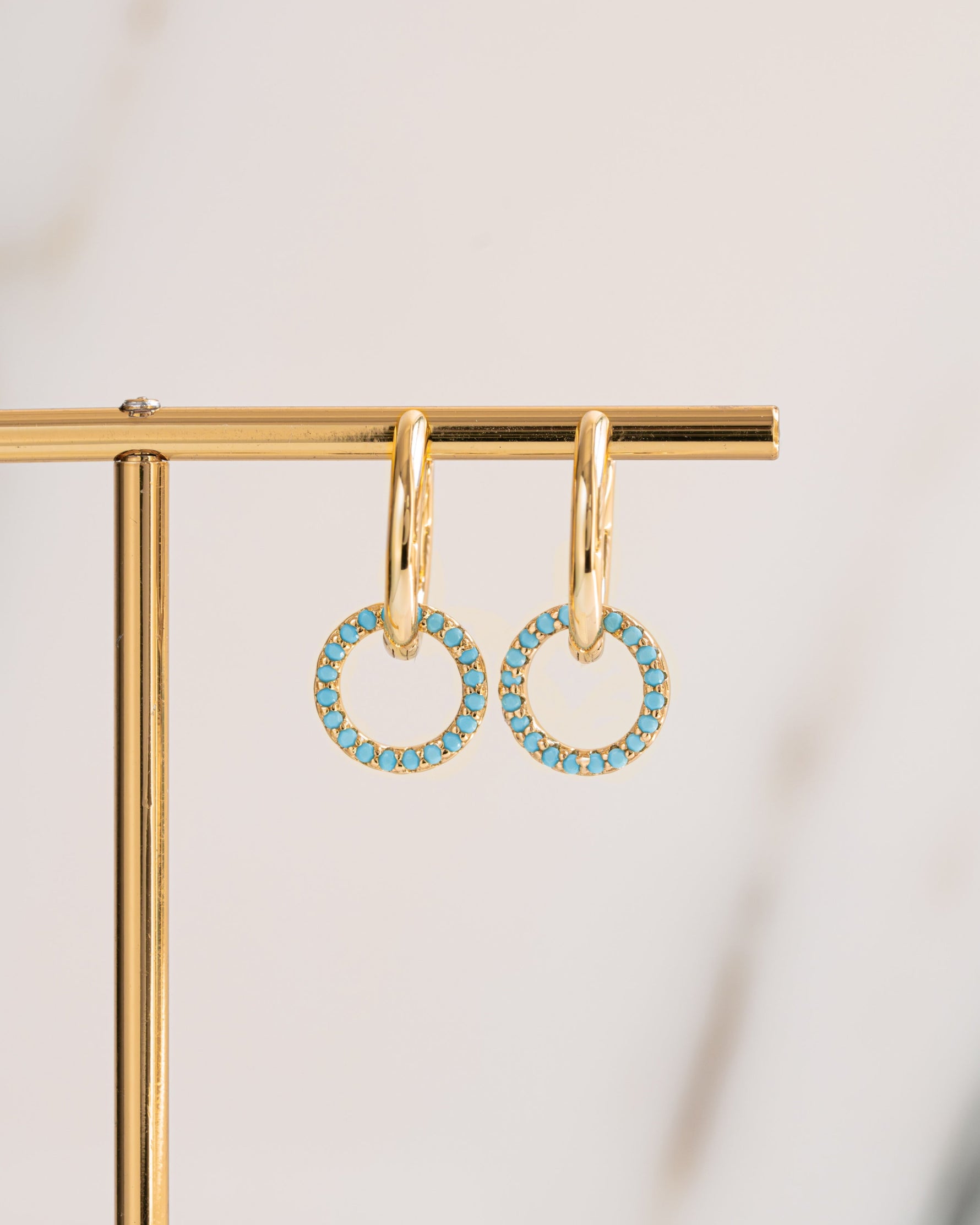 Small gold charm hoop earrings with turquoise gemstones on a gold stand against a light background