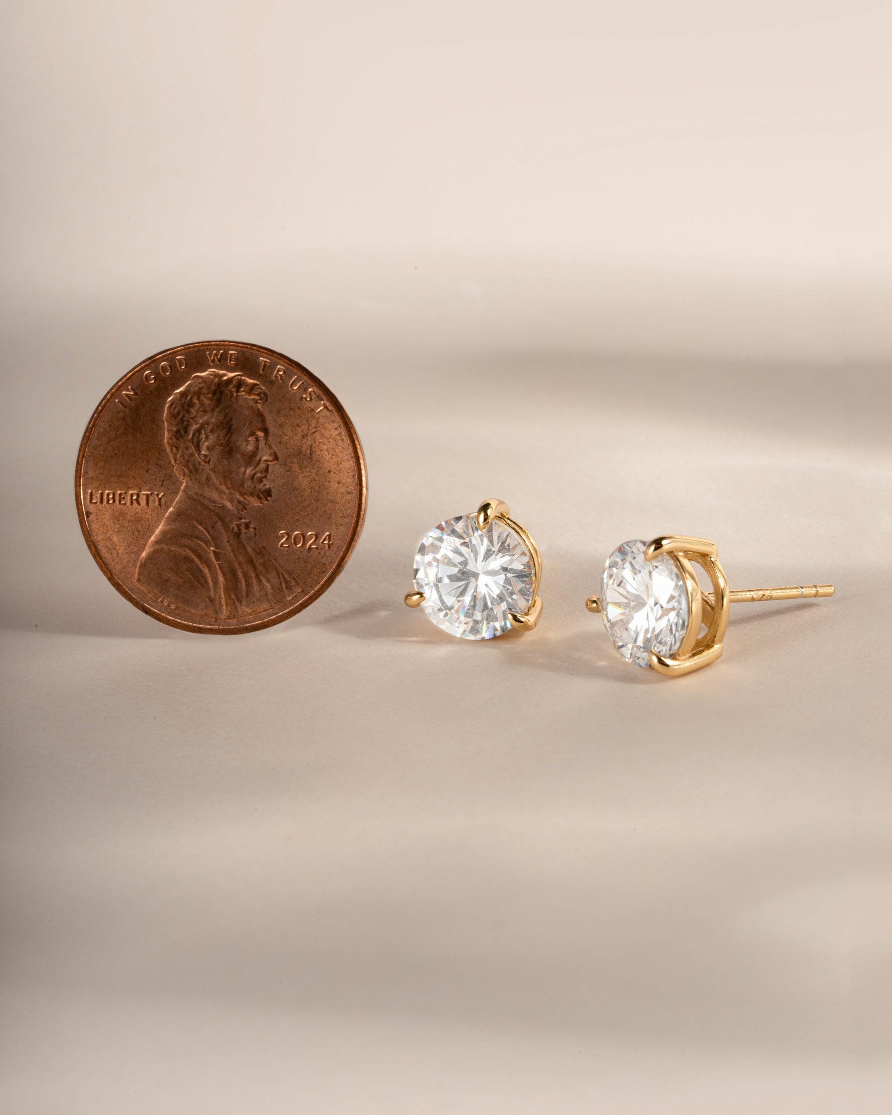 Gold studs being compared to the size of a penny on a beige background