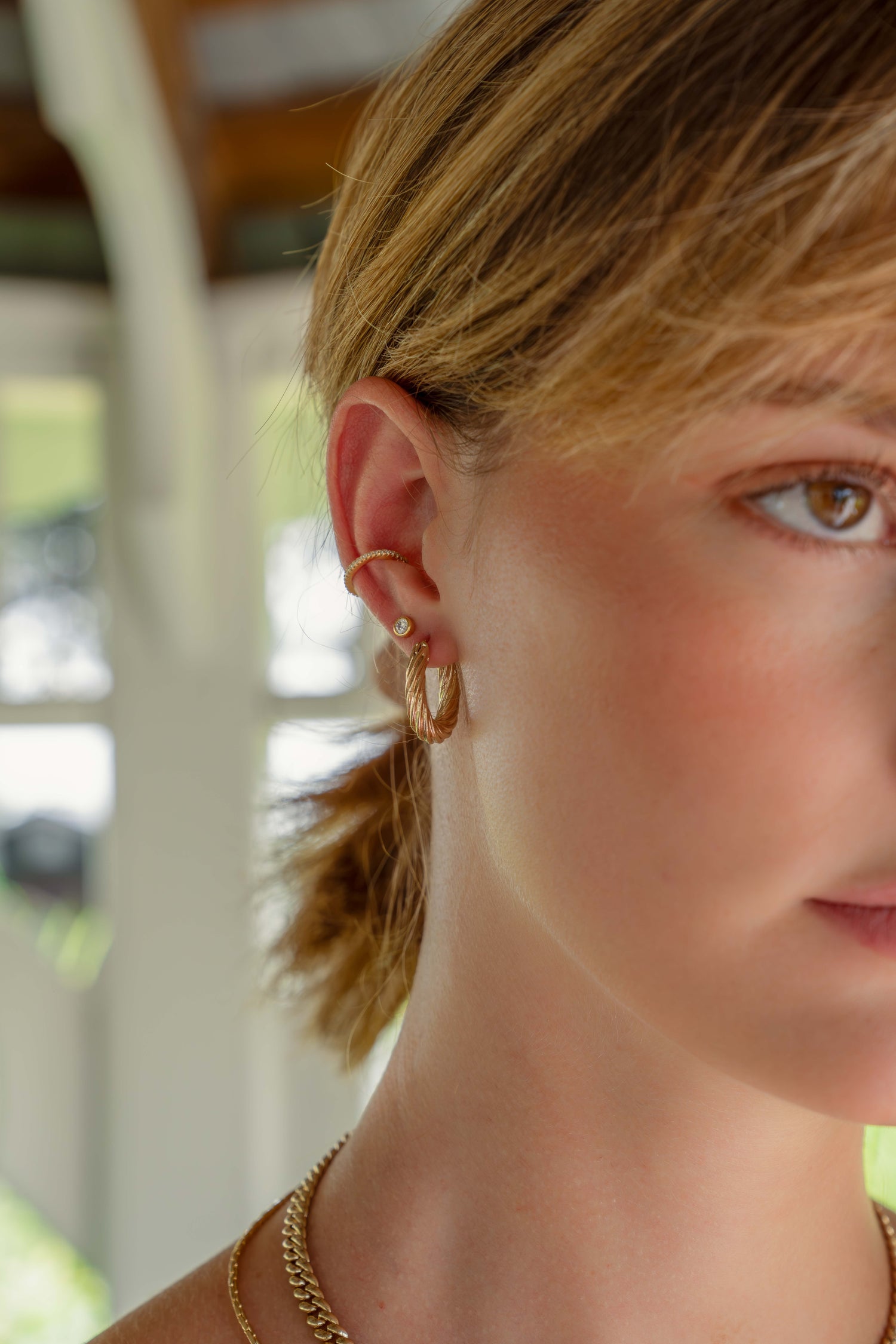 Model wearing gold earrings featuring a single cubic zirconia ear cuff, a bezel set stud earring and a gold textured hoop