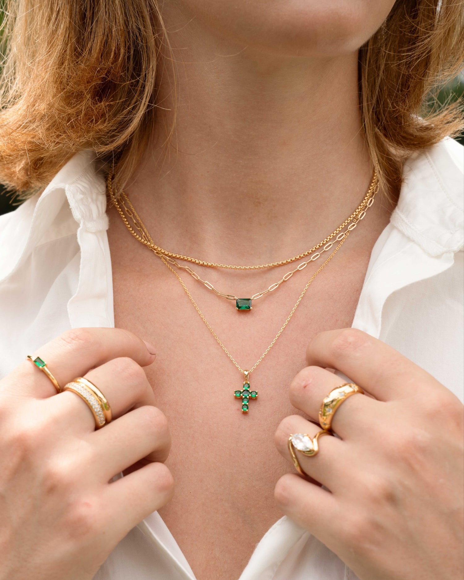 Model wearing emerald green layering necklaces and gold stackable rings featuring an emerald green emerald cut pendant necklace, and emerald cross necklace, and a gold chain layering necklace