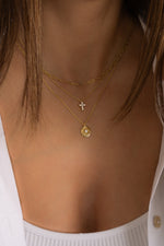 Model wearing dainty everyday gold necklaces in a stack featuring a dainty paperclip chain, a cross pendant, and a sunburst medallion pendant necklace. 
