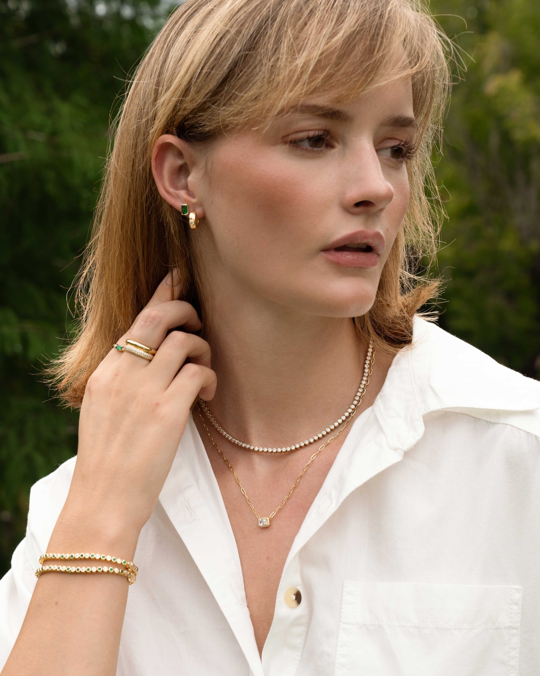 model wearing gold dainty jewelry featuring a tennis necklace, a baguette gold chain necklace, dainty emerald green stacking bracelets, and dainty gold rings