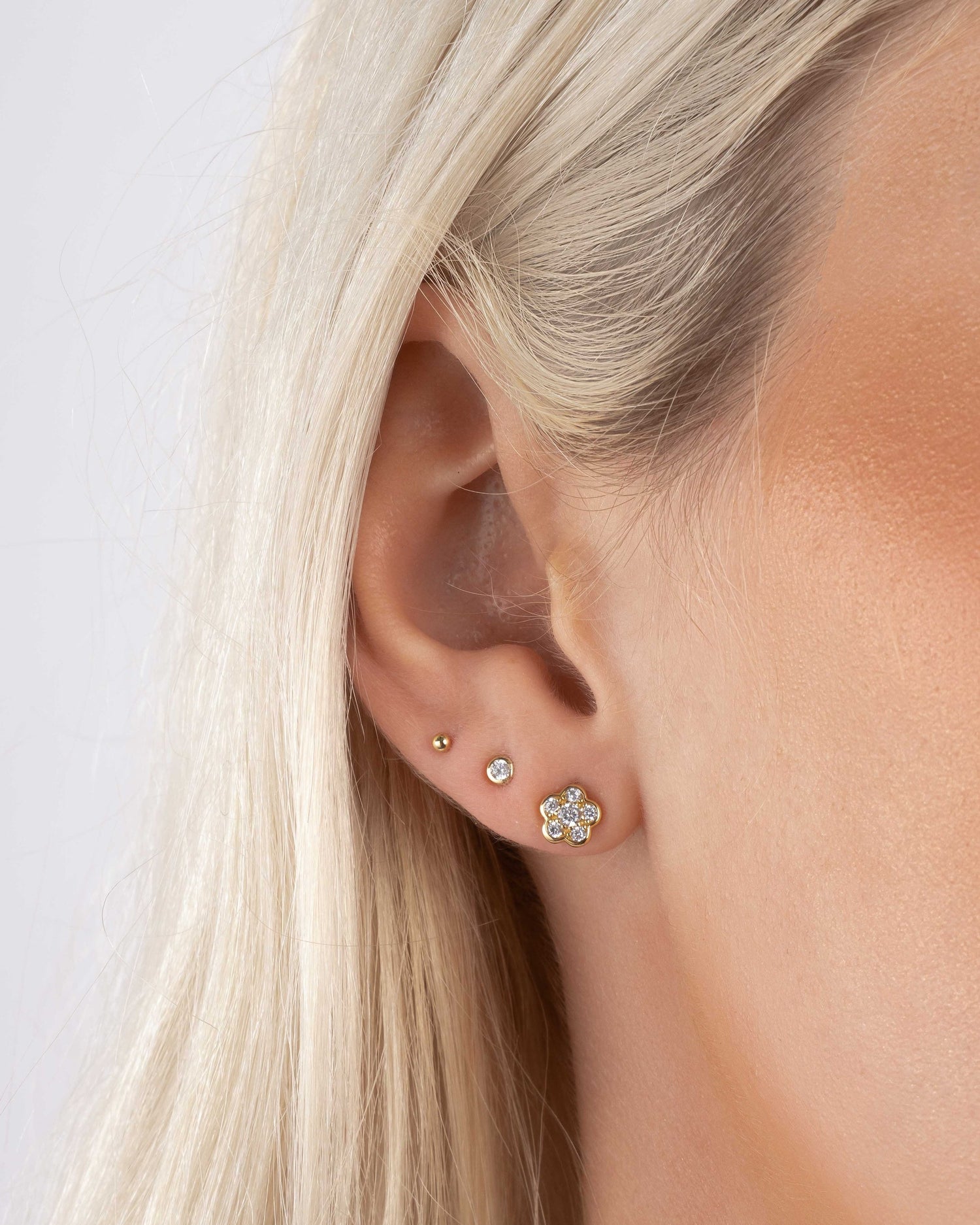 Closeup of a model wearing gold flower earrings, a diamond stud earring, and a dainty gold tiny earring. 