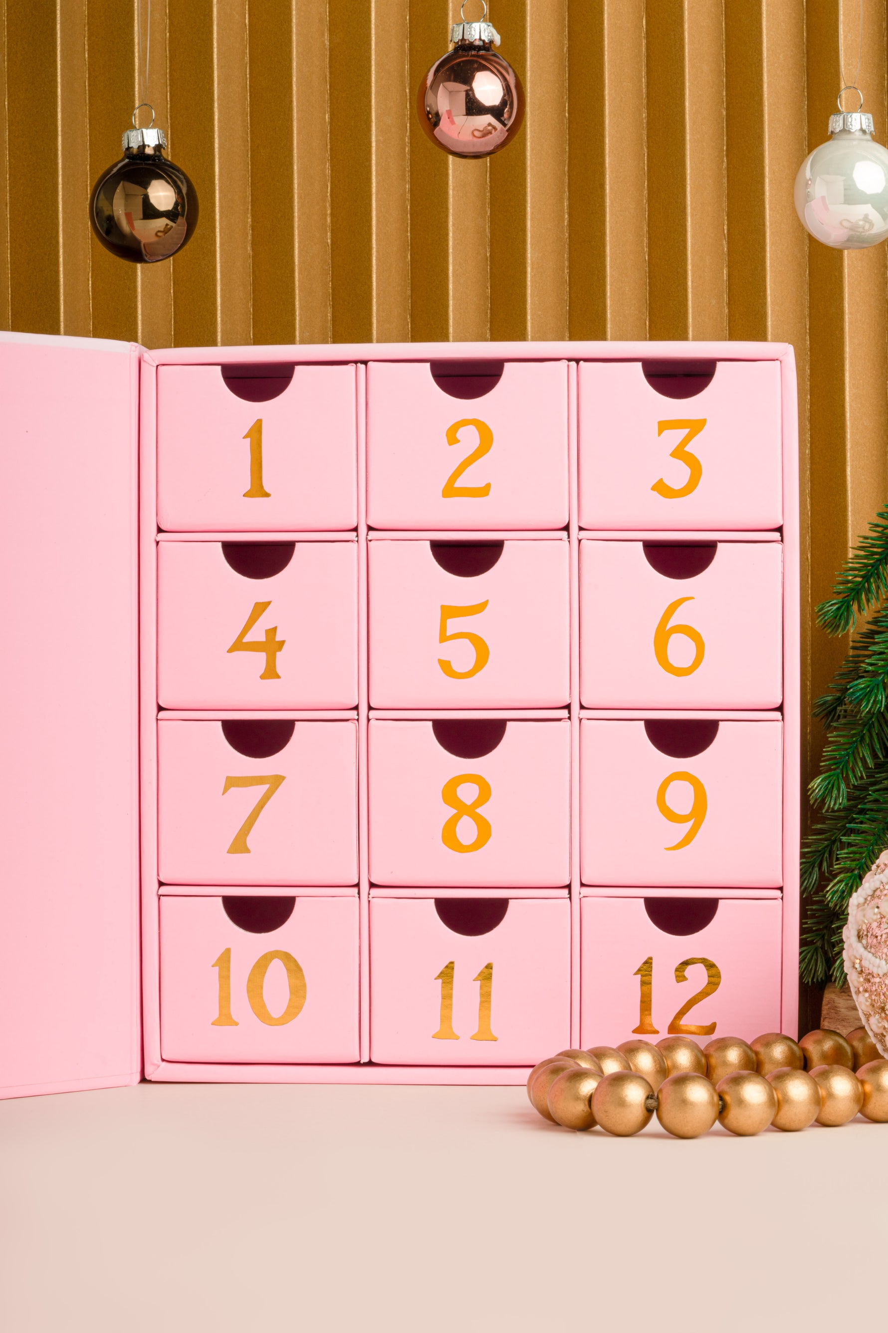 Pink advent calendar with gold numbers against a gold curtain background holding gold dainty jewelry, the perfect gift for her
