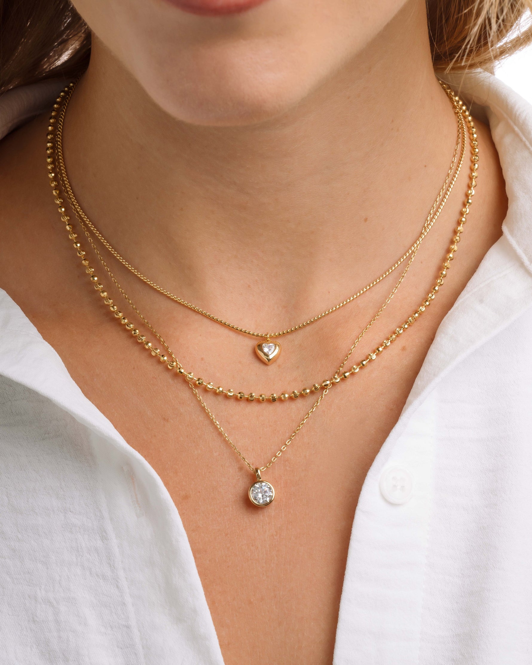Close-up of a person wearing gold layering necklaces, featuring a heart necklace, a gold chain and a round pendant necklace 
