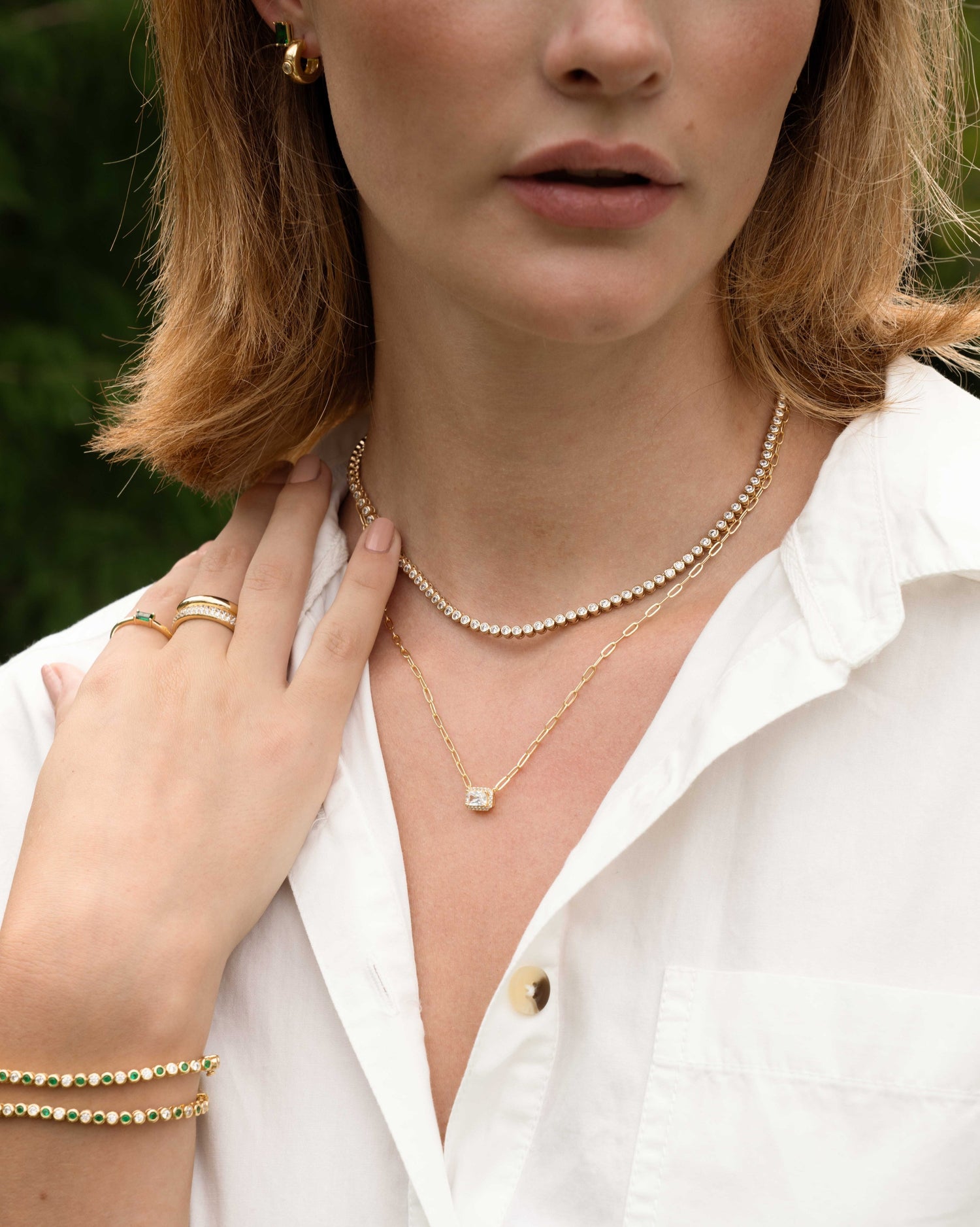 model wearing gold jewelry featuring a diamond tennis necklace, a gold pendant necklace, two emerald tennis bracelets, and gold rings