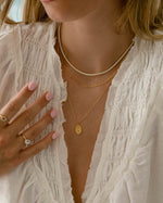Model wearing gold dainty necklaces, featuring a diamond tennis necklace, a gold chain necklace, and a oval medallion pendant necklace, with two diamond ringss