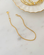 Gold chain bracelet on a marble surface with a decorative dish in the background.