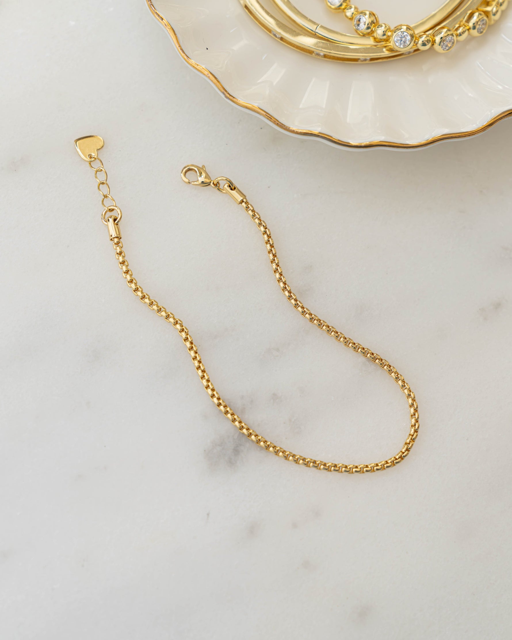 Gold chain bracelet on a marble surface with a decorative dish in the background.