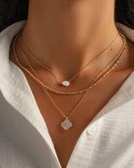 Gold layering necklace stack featuring a mother of pearl clover necklace, a pear diamond necklace, and a gold beaded chain necklace 