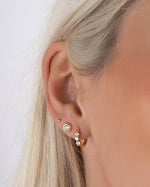 Close up image of models ear wearing gold small hoop earrings a diamond bezel stud earring, and a tiny earring ball stud 