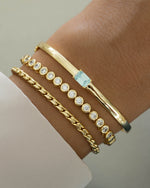 Gold bracelet stack featuring a aquamarine bracelet with a bangle design, a gold tennis bracelet, and a  gold chain bracelet on models wrist