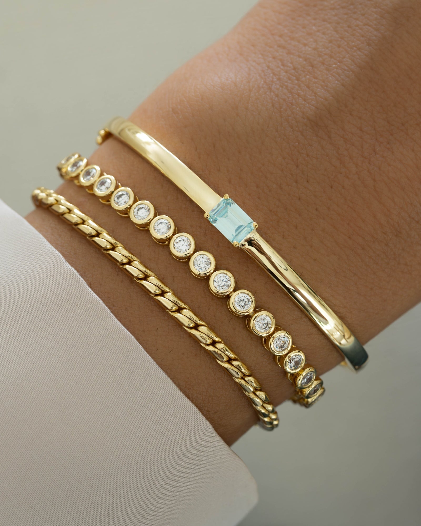 Gold bracelet stack featuring a aquamarine bracelet with a bangle design, a gold tennis bracelet, and a  gold chain bracelet on models wrist