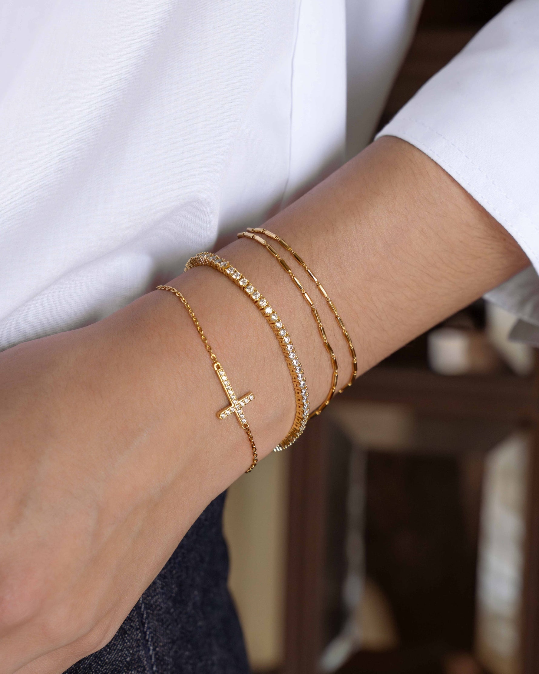 Closeup image of model wearing a gold bracelet stack featuring a dainty gold chain bracelet set, a diamond tennis bracelet, and a cross bracelet