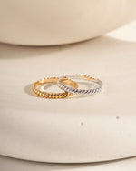 Two rings featuring a rope ring design in gold and sterling silver on a textured beige surface