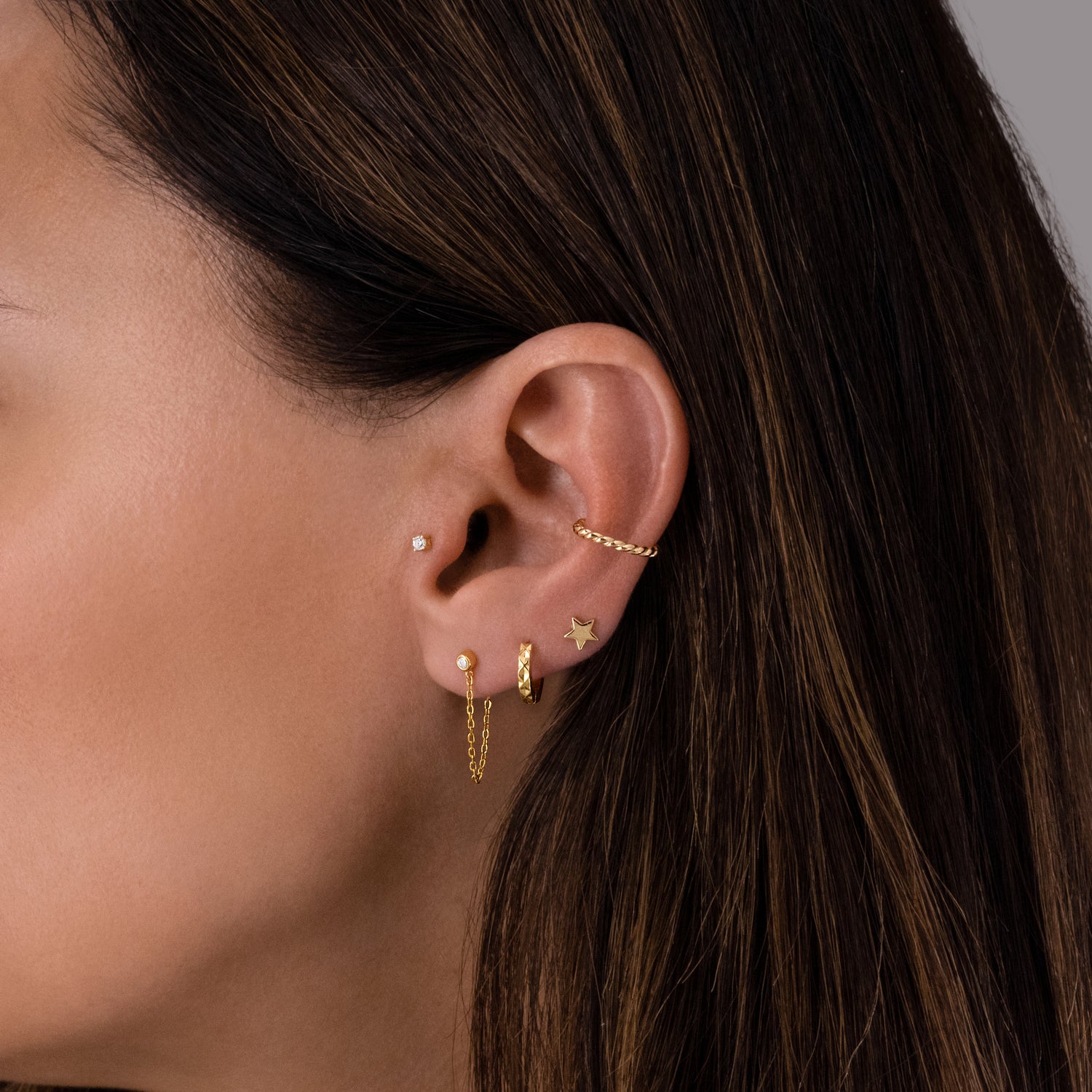 Model wearing gold earring stack featuring a diamond chain stud earrings, textured plain huggies, and a plain star stud earring. Model is also wearing a tragus stud earring and a minimalist spiral ear cuff.