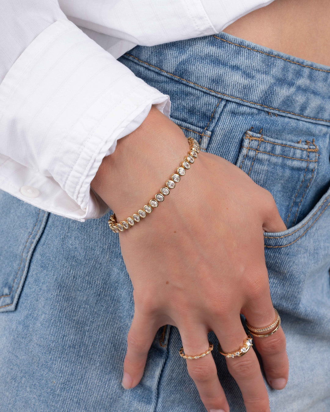 model wearing gold oval bezel set diamond tennis bracelet, and dainty gold stacking rings while hand is in pocket