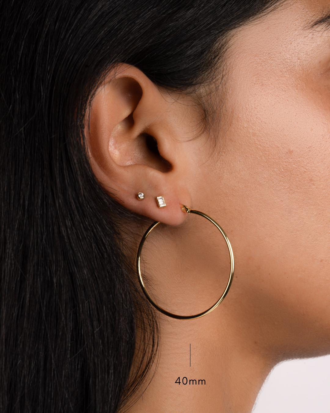 model wearing 40mm gold hoop earrings, a gold baguette stud, and a small gold bezel stud earring