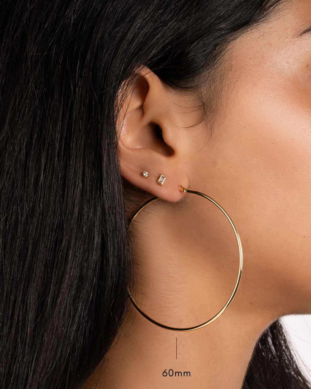 model wearing gold 60mm hoop earrings, a gold baguette stud, and a  gold small bezel stud earring