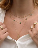 Model wearing gold and emerald layering necklaces and stacking rings, featuring a small paperclip chain necklace in gold, a beaded emerald flower necklace and a oval medallion pendant necklace
