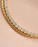 Gold bracelet stack with a rope chain bracelet and a tennis bracelet on a neutral background